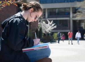 Female student sitting cross-legged in the Plaza writing in a notebook