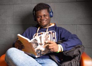 Smiling student sitting in a chair wearing headphones and holding a book